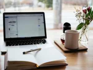 a desk with a mug on the right, an open laptop in the back, and a notebook opened to the middle with a pen sitting on top in the front of the desk.