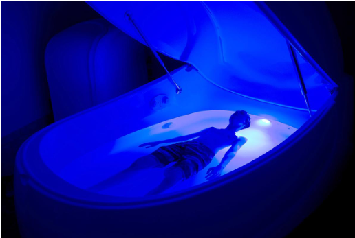 Person floating in float tank with open top and blue lights.